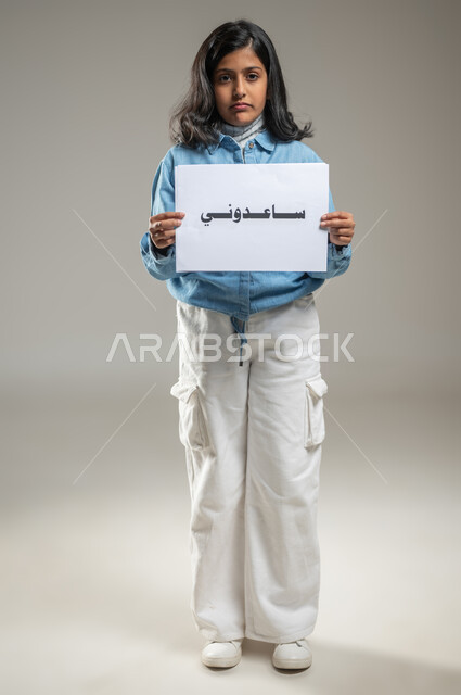 Awareness campaigns to reduce violence against children, the need to protect children and provide them with security, portrait of a Saudi Arabian Gulf girl holding in her hands a whiteboard with the words Help Me, looking at the camera with sad expressions, full-length body, gray background, sensitive content, sensitive use