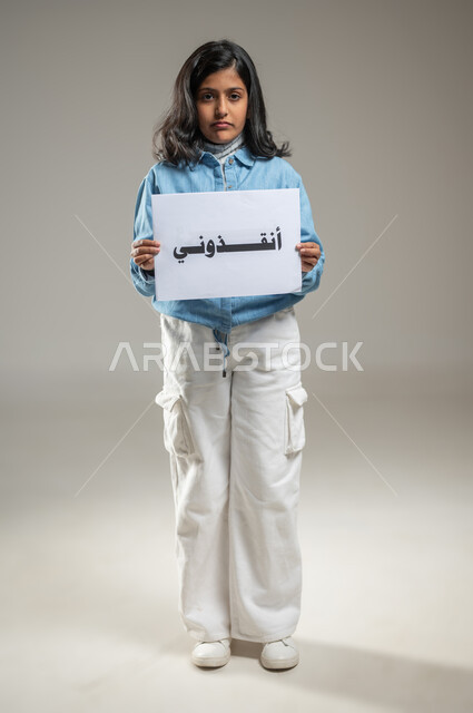 حماية الأطفال وتوفير ملاذ آمن لهم واجب على كل شخص في المجتمع, بورتريه لطفلة خليجية عربية سعودية تمسك بيديها ورقة مكتوب عليها أنقذوني, النظر إلى الكاميرا بتعبيرات الحزن, فتاة صغيرة تشعر بالخوف وتطلب النجدة, جسم بالطول الكامل, خلفية رمادية, محتوى حساس, استخدام حساس