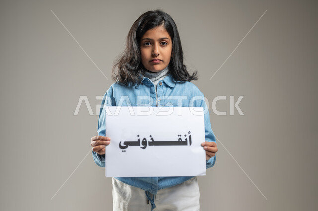 طلب المساعدة والإنقاذ, ضرورة حماية الأطفال وتوفير ملاذ آمن لهم, بورتريه لطفلة خليجية عربية سعودية تحمل ورقة بيضاء مكتوب عليها بالخط العريض أنقذوني, النظر إلى الكاميرا بتعبيرات الخوف والألم, فتاة تطلب النجدة, خلفية رمادية, محتوى حساس, استخدام حساس