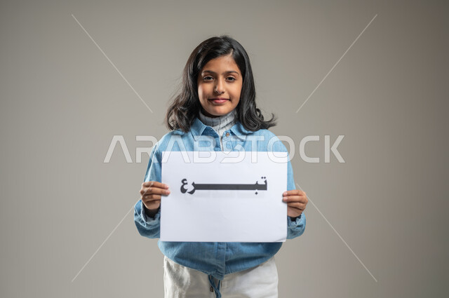 Urging people in society to engage in charitable work, launching awareness campaigns about the necessity of helping the poor and needy, portrait of a Saudi Gulf Arab girl holding a white board with a donation written on it, gestures of joy and happiness, gray background, sensitive content, sensitive use