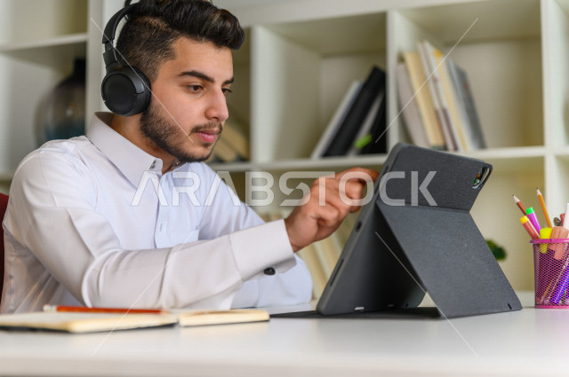 شاب سعودي خليجي يدرس في المنزل باستخدام جهاز التابلت، يتابع حصص الاونلاين ، التعلم عن بعد ، مفهوم التعليم عبر الانترنت ،الدراسة من خلال منصة تعليمية للتعلم عن بعد  ، العزلة الذاتية .