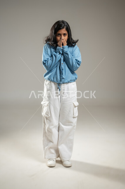 النظر إلى الكاميرا ووضع اليدين على الفم بإيماءات تدل على الدهشة والمفاجأة, بورتريه لفتاة عربية خليجة سعودية ترتدي زي كاجوال, الوقوف والتعبير عن المشاعر بحركات جسدية, جسم بالطول الكامل, خلفية رمادية, محتوى حساس
