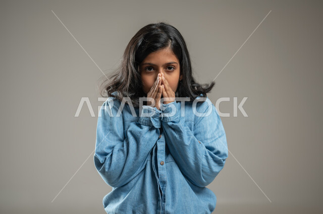 التعبير عن المشاعر بحركات جسدية, النظر إلى الكاميرا ووضع اليدين على الفم  وتغطيته بإيماءات تدل على الاندهاش والمفاجأة, بورتريه لفتاة عربية خليجة سعودية ترتدي قميص جميل لونه أزرق, خلفية رمادية, محتوى حساس