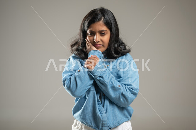 الإحساس بألم في الأسنان, وجوب الذهاب إلى طبيب الاسنان من أجل العلاج, بورتريه لبنت عربية خليجة سعودية ترتدي قميص أزرق اللون تضع يدها مكان آلامها , النظر إلى الكامبرا بتعبيرات التألم والانزعاج الشديد, خلفية رمادية, محتوى حساس