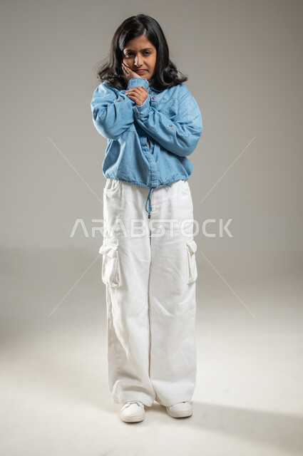 وضع اليد على الخد في إشارة إلى وجود ألم في الأسنان, بورتريه لفتاة عربية خليجة سعودية ترتدي زي كاجوال جميل تقف وتنظر إلى الكاميرا بإيماءات تدل على الأوجاع و الاستياء, الحاجة لزيارة طبيب الأسنان, جسم بالطول الكامل, محتوى حساس
