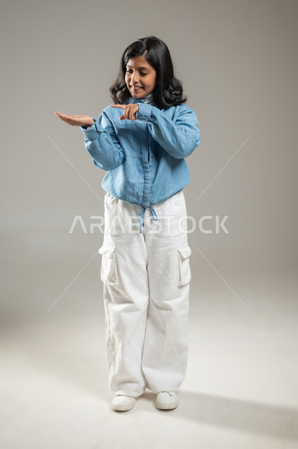 المتعة في اكتشاف الأشياء من حولنا , بورتريه لفتاة أنيقة عربية خليجة سعودية ترتدي زي كاجوال جميل تنظر إلى يدها, الوقوف والإشارة إلى شيء ما موجود في كفها الصغيرة, جسم بالطول الكامل, خلفية رمادية, محتوى حساس