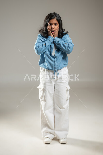 الوقوف والنظر إلى الكاميرا بشكل مباشر ووضع الكفين على الوجه, إيماءات تدل على المفاجأة والدهشة, بورتريه لفتاة لطيفة عربية خليجة سعودية تلبس قميص أزرق جميل وبنطال أبيض اللون, جسم بالطول الكامل, خلفية رمادية, محتوى حساس