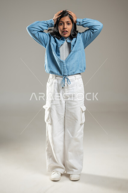 Looking at the camera and placing hands on the head, gestures indicating getting into trouble, portrait of a cute Saudi Arabian girl wearing a beautiful blue shirt and white pants, full-length body, gray background, sensitive content