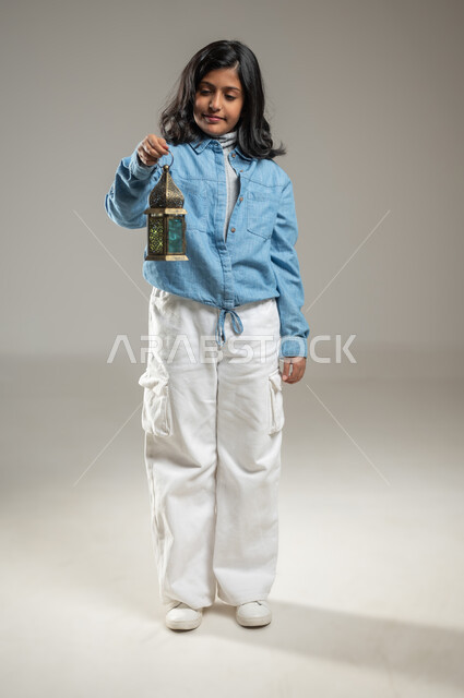 Celebrating the advent of the holy month of Ramadan, a portrait with a gray background of a Saudi Gulf Arab girl holding a beautiful ornate metal lantern in her hand, an elegant girl wearing blue and white, a full-length body, looking at the lantern with gestures of joy and happiness, decorations for Islamic holidays and occasions, sensitive content