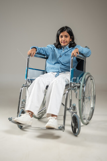 التعامل مع الأطفال ذوي الاحتياجات الخاصة بايجابية وتفاؤل, بورتريه لفتاة عربية خليجة سعودية تلبس قميص أزرق وبنطال أبيض اللون مقعدة تجلس على كرسي متحرك, النظر إلى الكاميرا بإيماءات السعادة والأمل, طفلة من ذوي الهمم لطيفة أنيقة، خلفية رمادية, محتوى حساس