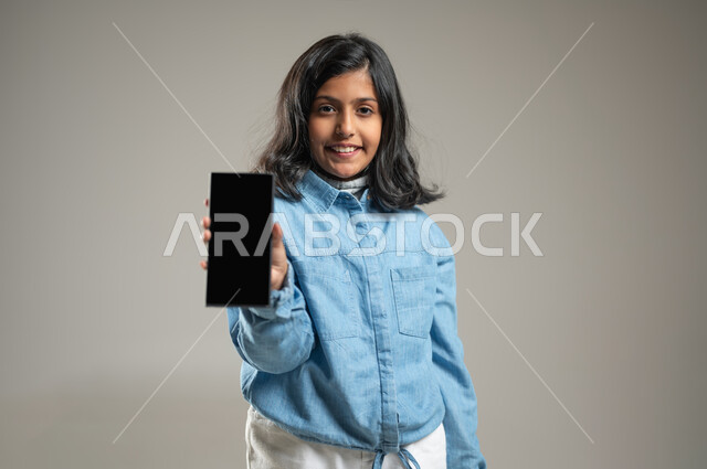 تطوير مستقبل الأطفال في المملكة العربية السعودية بالاستفادة من الأجهزة الذكية, بورتريه لفتاة لطيفة عربية خليجة سعودية تلبس قميص أزرق جميل, تحمل بيدها الجوال, عرض شاشة سوداء للهاتف المحمول, النظر للكاميرا مبتمسة, خلفية رمادية, محتوى حساس