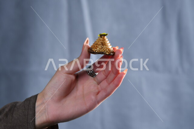 تقديم الحلويات والسكريات في الأعياد والمناسبات في السعودية، صورة مقربة ليد امرأة عربية تمسك بقطعة من الحلوى، تذوق ضيافة العيد، حلا عربي شرقي، خلفية رمادية