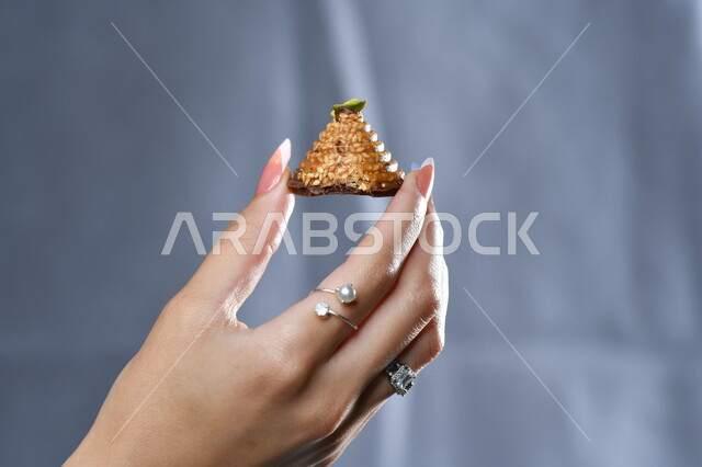 تذوق ضيافة العيد، تقديم الحلويات والسكريات في الأعياد والمناسبات في السعودية، صورة مقربة ليد امرأة عربية تمسك بقطعة من الحلوى، حلا عربي شرقي، خلفية رمادية