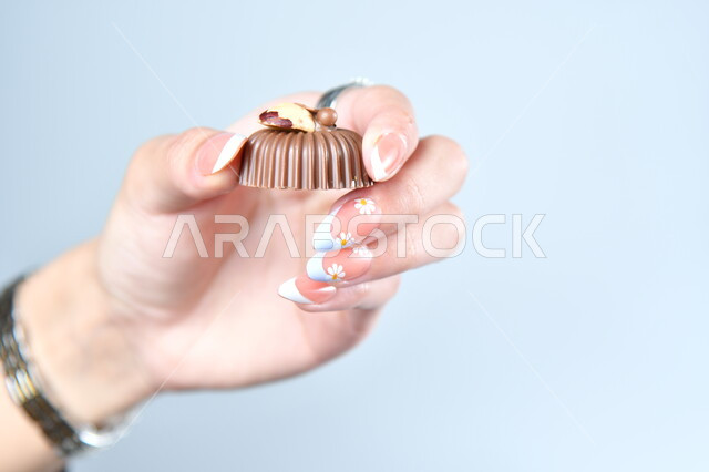 أنواع وأصناف الشوكولاته الفاخرة، تقديم الحلويات والسكريات في الأعياد والمناسبات في السعودية، صورة مقربة ليد امرأة عربية تمسك بقطعة من الشوكولا، حلا عربي شرقي، خلفية رمادية