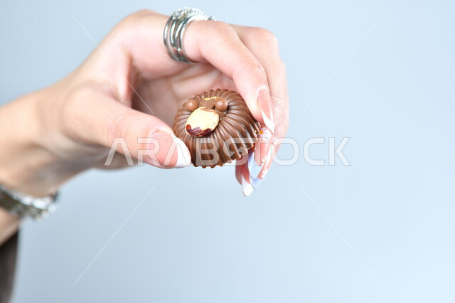 الأشكال الفنية المختلفة لحبات الشوكولاته الفاخرة، تقديم الحلويات والسكريات في الأعياد والمناسبات في السعودية، صورة مقربة ليد امرأة عربية تمسك بقطعة من الشوكولا، حلا عربي شرقي، خلفية رمادية