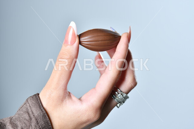 حبات الشوكولاته الفاخرة بأشكال فنية مختلفة، تقديم الحلويات في الأعياد والمناسبات، صورة مقربة ليد امرأة عربية تمسك بقطعة من الشوكولا، خلفية رمادية