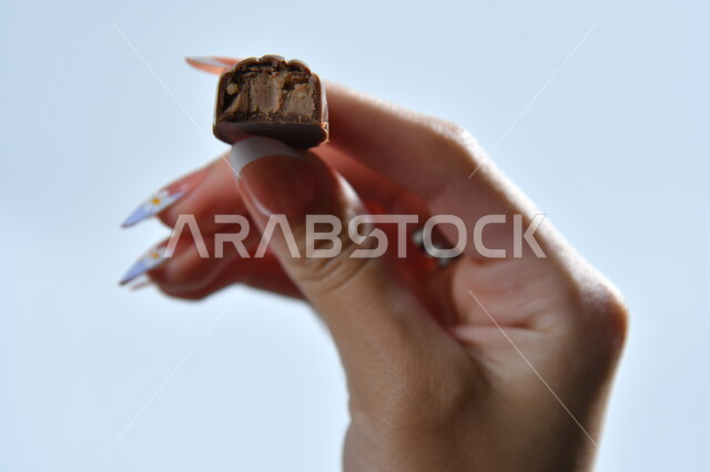 أنواع  الشوكولاته الفاخرة، تقديم الحلويات والسكريات في الأعياد والمناسبات، صورة مقربة ليد امرأة عربية تمسك بقطعة من الشوكولا، تذوق قطعة من الحلا، خلفية رمادية