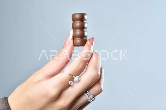 الأشكال الفنية المختلفة لحبات الشوكولاته الفاخرة، تقديم الحلويات في الأعياد والمناسبات، صورة مقربة ليد امرأة عربية تمسك بقطعة من الشوكولا، خلفية رمادية