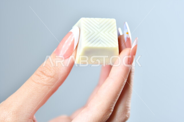 الشوكولاته البيضاء الفاخرة بأشكال فنية مختلفة، تقديم الحلويات في الأعياد والمناسبات، صورة مقربة ليد امرأة عربية تمسك بقطعة من الشوكولا، خلفية رمادية