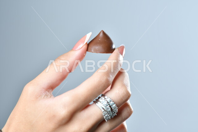 حبات الشوكولاته الفاخرة بأشكال فنية مختلفة، تقديم الحلويات في الأعياد والمناسبات، صورة مقربة ليد امرأة عربية تمسك بقطعة من الشوكولا، خلفية رمادية