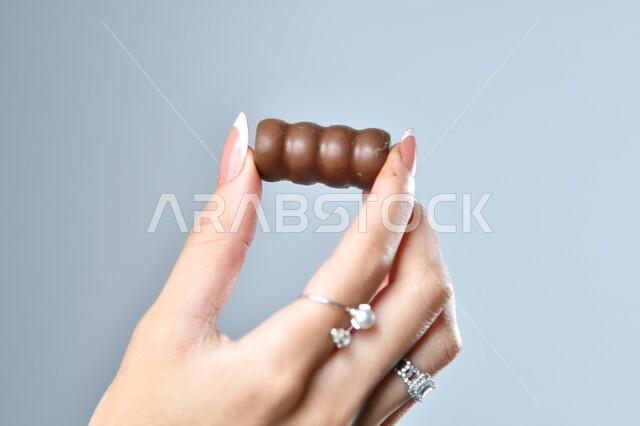 ضيافة العيد السعيد، تقديم الحلويات في الأعياد والمناسبات، الأشكال الفنية المختلفة لحبات الشوكولاته الفاخرة، صورة مقربة ليد امرأة عربية تمسك بقطعة من الشوكولا، خلفية رمادية