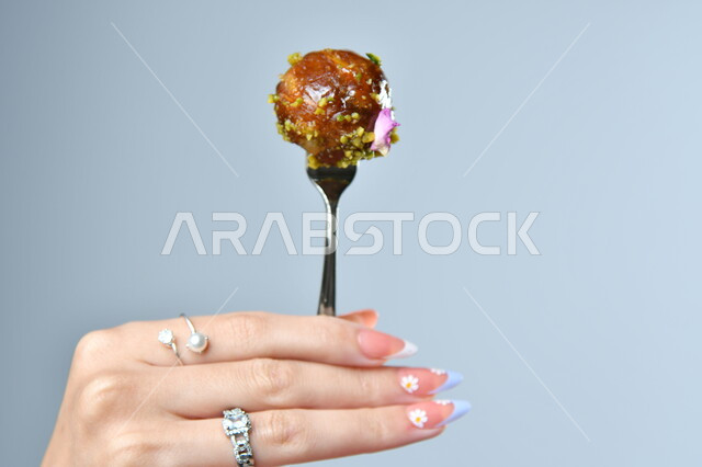 Eastern Arab food, popular Arabic sweets, a close-up of an Arab woman’s hand holding a fork containing a piece of delicious luqaimat, high-calorie foods, gray background