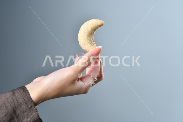 بسكويت على شكل هلال، تقديم الحلويات والسكريات في الأعياد والمناسبات في السعودية، صورة مقربة ليد امرأة عربية تمسك بقطعة من الحلوى، حلا عربي شرقي، خلفية رمادية