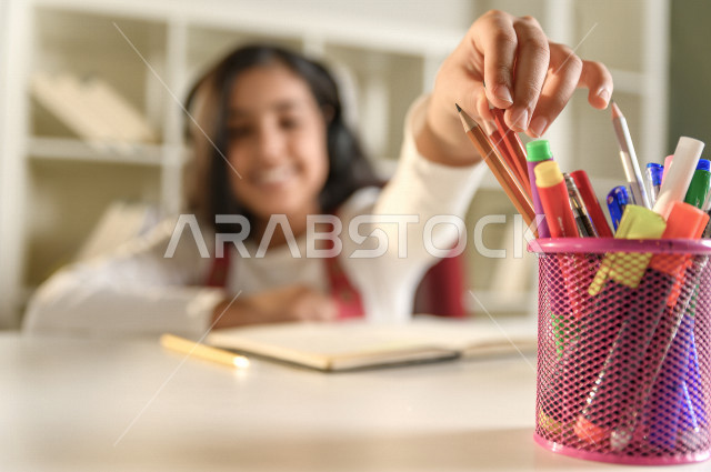 تلميذة سعودية خليجية سعيدة بالدراسة في المنزل ، تختار قلم مناسب لتأدية الواجب المدرسي  ،الدراسة من خلال منصة تعليمية للتعلم عن بعد ،  التعلم عن بعد ، مفهوم التعليم عبر الانترنت ، التعليم المنزلي ، العزلة الذاتية .