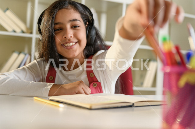 تلميذة سعودية خليجية سعيدة بالدراسة في المنزل ، تختار قلم مناسب لتأدية الواجب المدرسي  ،الدراسة من خلال منصة تعليمية للتعلم عن بعد ،  التعلم عن بعد ، مفهوم التعليم عبر الانترنت ، التعليم المنزلي ، العزلة الذاتية .