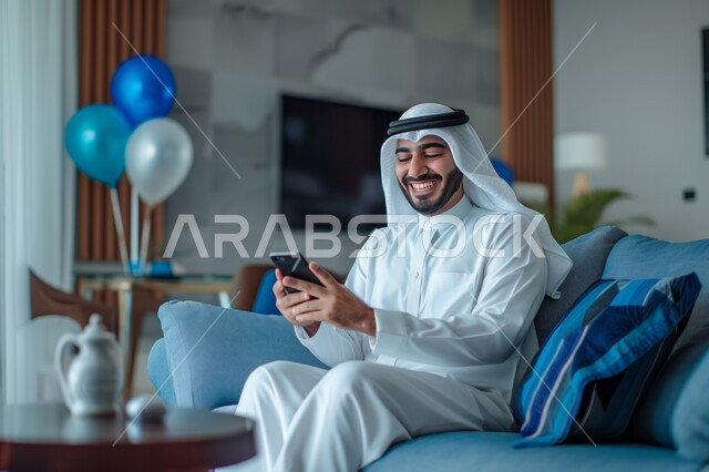 Expressions and gestures of joy and happiness, a Saudi Gulf Arab man wearing a ghutra and traditional dress holding a mobile phone in his hand, sitting in the living room, spending enjoyable times at home, using modern technical and technological devices, communicating and chatting with friends and relatives via mobile phone.