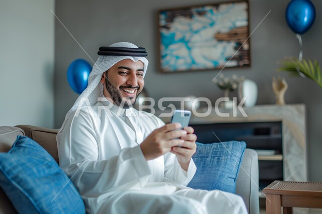 Spending enjoyable times at home, using modern technical and technological devices, communicating and chatting with friends and relatives via mobile phone, expressions and gestures of joy and happiness, a Saudi Gulf Arab man wearing a ghutra and traditional dress, holding a mobile phone in his hand, sitting in the living room.