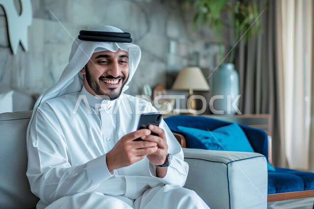 Using modern technical and technological devices, communicating and chatting with friends and relatives via mobile phone, expressions and gestures of joy and happiness, a Saudi Gulf Arab man wearing a ghutra and traditional dress holding a mobile phone in his hand sitting in the living room, spending enjoyable times at home