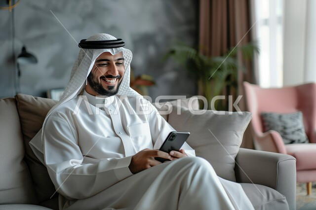 Communicating and chatting with friends and relatives via mobile phone, expressions and gestures of joy and happiness, a Saudi Gulf Arab man wearing a ghutra and traditional dress holding a mobile phone in his hand sitting in the living room, spending enjoyable times at home, using modern technical and technological devices