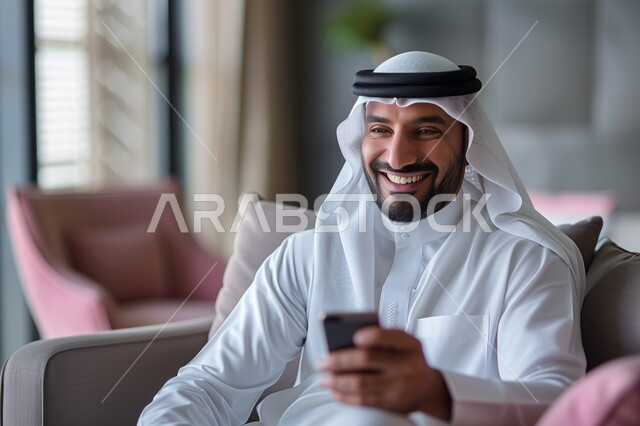 Gestures and expressions of joy and happiness, a Saudi Gulf Arab man wearing a ghutra and traditional dress holding a mobile phone in his hand, sitting in the living room, spending enjoyable times at home, using modern technical and technological devices, communicating and chatting with friends and relatives via mobile phone
