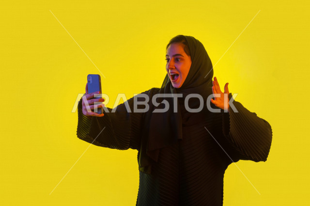 بورتريه شابة سعودية بايماءات وجه و يدين تدل على الفرحة و الحيوية ، مصورة خليجية تحمل هاتفها المحمول ، تلتقط صور سلفي  ، خلفية ملونة