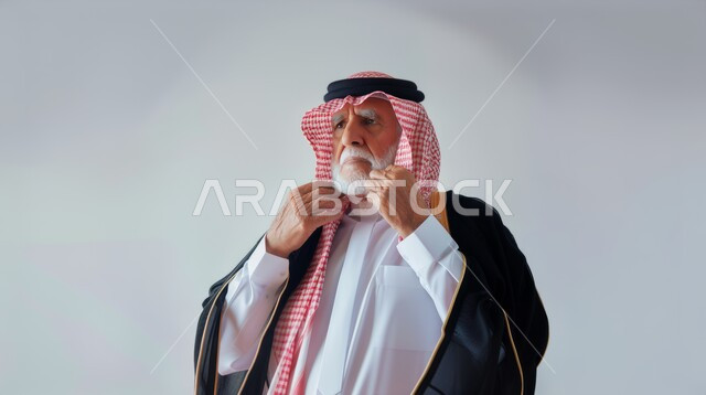 تعابير وايماءات الرجولة والثقة بالنفس، بورتريه مقرب لرجل كبير بالسن عربي خليجي سعودي يرتدي الثوب والبشت التقليدي يقوم بتعديل شماغه، ارتداء الأزياء الشعبية السعودية، الاهتمام بالأناقة والترتيب والمظهر الخارجي، خلفية رمادية