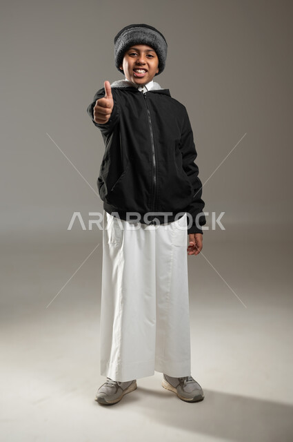 الموافقة على أمر معين، بورتريه لولد عربي خليجي سعودي مبتسم يرتدي الثوب التقليدي مع ملابس الشتاء الدافئة يرفع اصبع الإبهام للأعلى بتعابير علامة الجودة، النظر إلى الكاميرا بايماءات الإعجاب والرضا، الجسم بالطول الكامل، خلفية رمادية، محتوى حساس، استخدام حساس