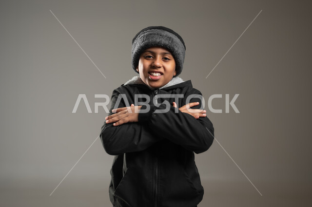 ملابس مناسبة لفصل الشتاء، النظر الى الكاميرا بايماءات وجه ويدين تدل على البرد الشديد، بورتريه مقرب لولد عربي خليجي يرتدي الثوب التقليدي و ملابس الشتاء ويضع يديه على كتفه بشكل متقاطع، خلفية رمادية, محتوى حساس , استخدام حساس