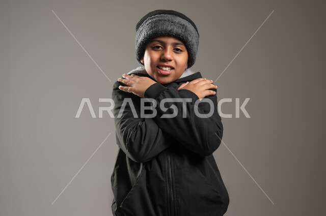 الأجواء الشتوية في السعودية، النظر الى الكاميرا بايماءات وجه ويدين تدل على البرد الشديد، ملابس مناسبة لفصل الشتاء، بورتريه مقرب لولد عربي خليجي يرتدي الثوب التقليدي و ملابس الشتاء ويضع يديه على كتفه بشكل متقاطع، خلفية رمادية, محتوى حساس , استخدام حساس