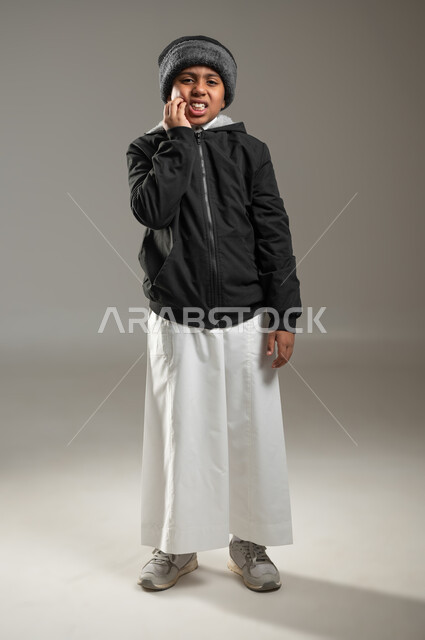 الحاجة لزيارة طبيب الأسنان، إلتهاب اللثة وتسوس الأسنان، بورتريه لولد عربي خليجي سعودي يعاني من الم في الاضراس، ايماءات الوجه واليد تدل على الوجع والالام، التعب والإرهاق وإنتفاخ الفم، خلفية رمادية، الجسم بالطول الكامل، محتوى حساس، استخدام حساس