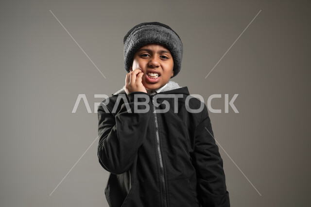 إلتهاب اللثة وتسوس الأسنان، الحاجة لزيارة طبيب الأسنان، بورتريه مقرب لولد عربي خليجي سعودي يعاني من الم في الاضراس، ايماءات الوجه واليد تدل على الوجع والالام، التعب والإرهاق وإنتفاخ الفم، خلفية رمادية، محتوى حساس، استخدام حساس