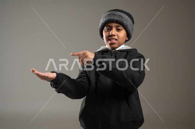 الاشارة بإصبع السبابة إلى شيء محدد, الوقوف والنظر إلى الكاميرا بإيماءات السرور,  بورتريه مقرب لولد عربي سعودي خليجي يرتدي الثوب التقليدي وملابس الشتاء الدافئة يحمل بيده شيء ما, رفع وحمل عنصر معين, خلفية رمادية، محتوى حساس، استخدام حساس