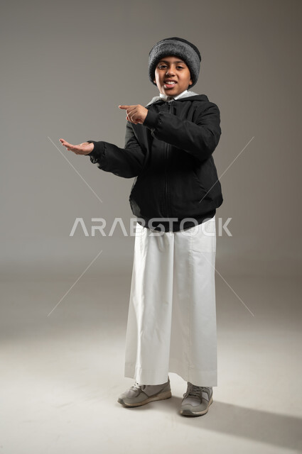 الوقوف والنظر إلى الكاميرا بإيماءات السرور, الاشارة بإصبع السبابة إلى شيء محدد, بورتريه لولد عربي سعودي خليجي يرتدي الثوب التقليدي وملابس الشتاء الدافئة يحمل بيده شيء ما, رفع وحمل عنصر معين, خلفية رمادية، الجسم بالطول الكامل، محتوى حساس، استخدام حساس