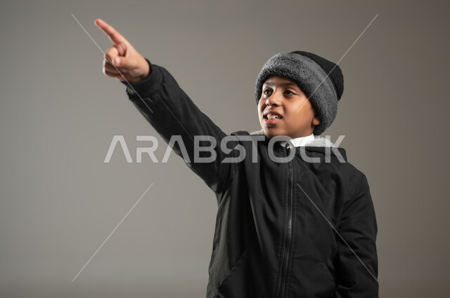 الوقوف والنظر إلى شيء ما, بورتريه مقرب لولد عربي سعودي خليجي يرتدي الثوب التقليدي وملابس الشتاء الدافئة يرفع اصبعة السبابة لجهة اليسار لتعين موقع محدد, الإشارة لمكان ما, خلفية رمادية، محتوى حساس، استخدام حساس