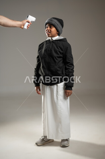 الحاجة إلى العلاج والمساعده، قياس درجة الحرارة بإستخدام الميزان الحراري، بورتريه لولد عربي سعودي خليجي يرتدي الثوب التقليدي وملابس الشتاء الدافئة يعاني من حمى شديدة، الإحساس والشعور بالمرض والإرهاق، الجسم بالطول الكامل، خلفية رمادية، محتوى حساس، استخدام حساس