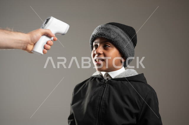 قياس درجة الحرارة بإستخدام الميزان الحراري، الحاجة إلى العلاج والمساعده، بورتريه مقرب لولد عربي سعودي خليجي يرتدي الثوب التقليدي وملابس الشتاء الدافئة يعاني من حمى شديدة، الإحساس والشعور بالمرض والإرهاق، خلفية رمادية، محتوى حساس، استخدام حساس