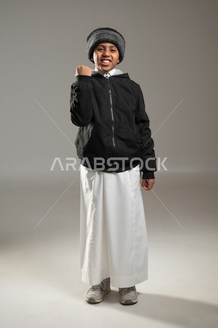 ايماءات تدل على الحماس والفرح، الفرح بالفوز والانتصار، بورتريه لولد عربي خليجي سعودي يرتدي الثوب التقليدي وملابس الشتاء الدافئة، النجاح والقوة وتحقيق الاهداف، خلفية رمادية، الجسم بالطول الكامل، محتوى حساس، استخدام حساس