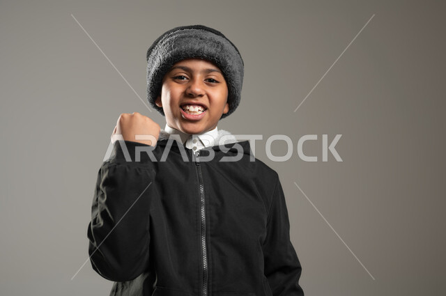 إيماءات وجه ورفع اليد تدل على الشغف والحماس، الفرح بالفوز والانتصار، بورتريه مقرب لولد عربي خليجي سعودي يرتدي الثوب التقليدي وملابس الشتاء الدافئة، النجاح والقوة وتحقيق الاهداف، خلفية رمادية، محتوى حساس، استخدام حساس
