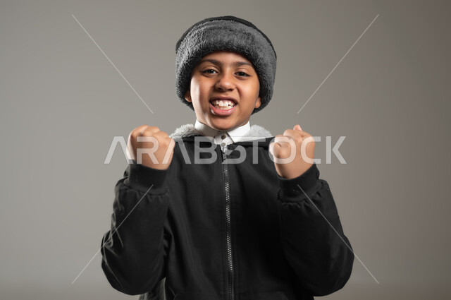 ايماءات وجه ويدين تدل على التشجيع والحماس، بورتريه مقرب لولد عربي خليجي سعودي يرتدي الثوب التقليدي وملابس الشتاء الدافئة، الفرح بالفوز والانتصار، النجاح والقوة وتحقيق الاهداف، خلفية رمادية، محتوى حساس، استخدام حساس