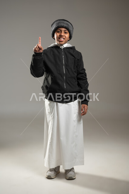 الوقوف باستقامة والنظر إلى الكاميرا, ايماءات الاستفسار والاشارة إلى شيء ما, بورتريه لولد عربي خليجي سعودي يرتدي الثوب التقليدي وملابس الشتاء الدافئة يرفع اصبعه للأعلى بإيماءات التوضيح, صورة الجسم بالطول الكامل , خلفية رمادية، محتوى حساس، استخدام حساس